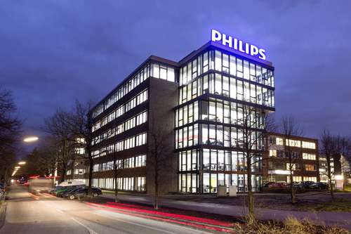 Foto: Mehrstöckiges Gebäude mit vielen beleuchteten Fenstern und einem leuchtenden Schriftzug PHILIPS auf dem Dach, unter dunkelblauen Abendhimmel. Ein beleuchtete Straße links im Vordergrund.
